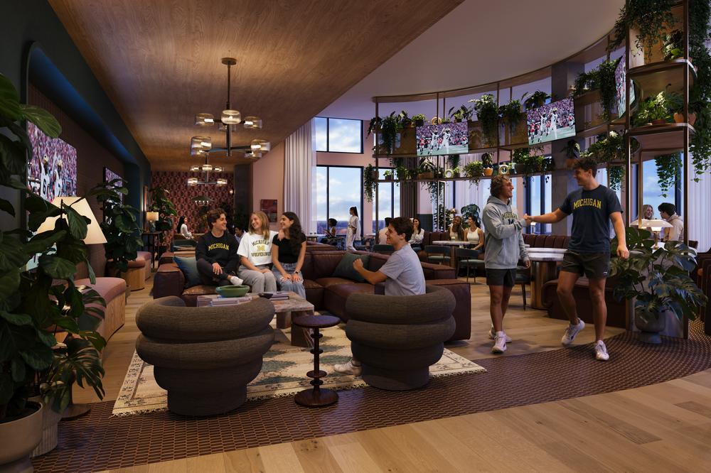 A group of young adults socialize in the modern, stylish Rambler Ann Arbor lounge, filled with plants, large windows, and comfortable seating. Two people greet each other while others sit and chat on sofas and chairs.