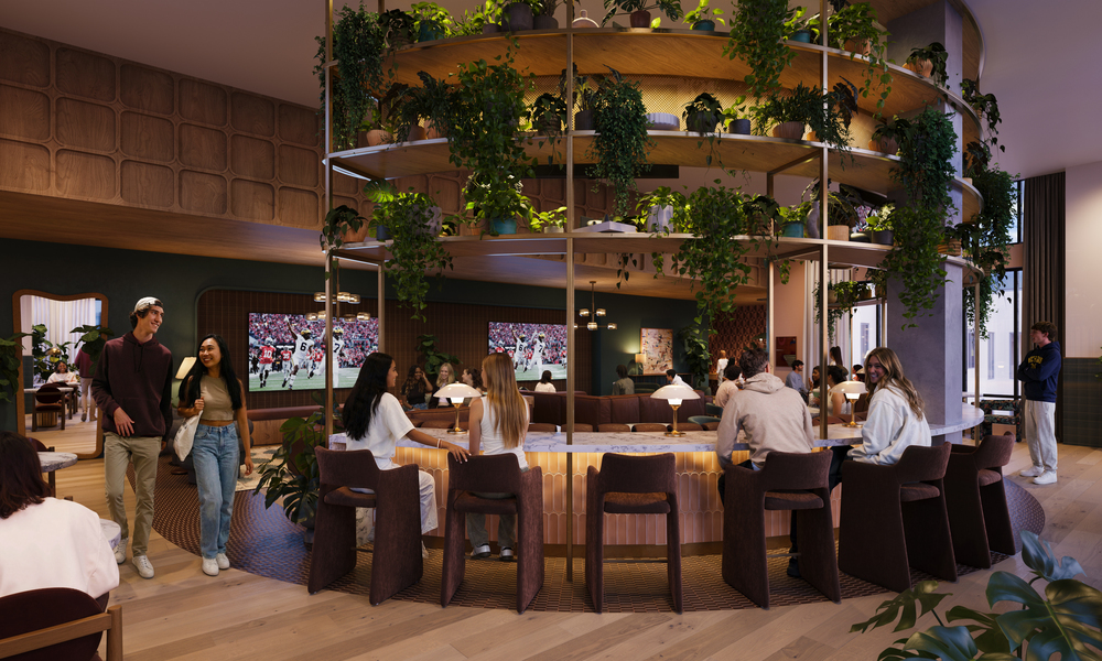 A modern lounge at Rambler Ann Arbor with people sitting and standing around a circular bar, surrounded by lush green plants, warm lighting, and wood accents, creating a lively and inviting atmosphere.
